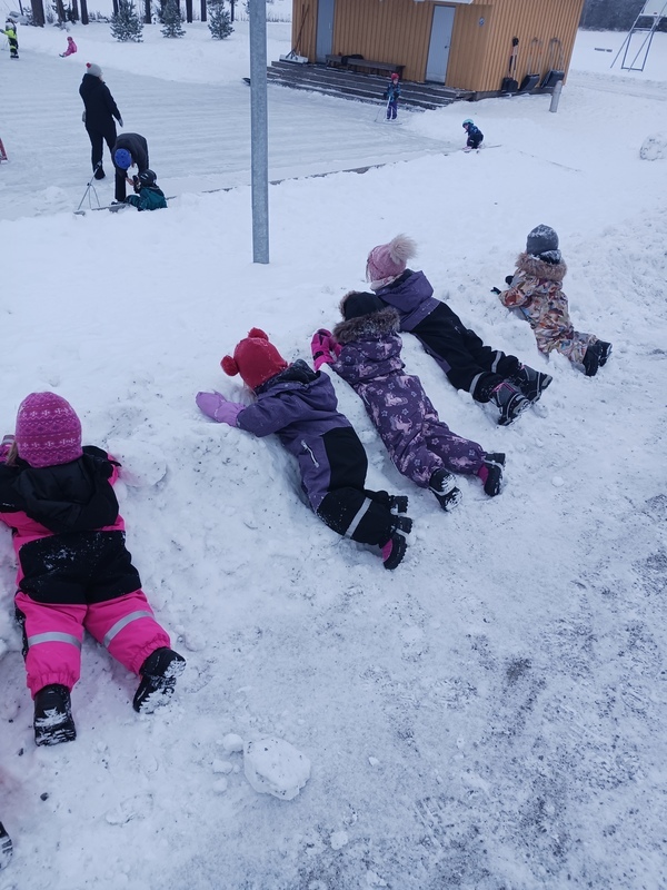 Lapset makaa lumihangessa ja katselee luistelijoita
