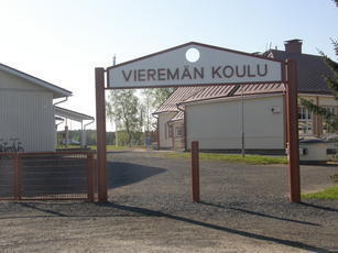 Kuvassa Vieremän koulu pihaportin luota katsottuna: punaisten portinpylväiden päällä on kolmiolevy, jossa lukee isoin kirjaimin Vieremän koulu. Portin läpi näkyy vanhan, puisen koulurakennuksen pääty ja vasemmalla näkyy uudemman koulurakennuksen päätyä. Aurinkoinen päivä.
