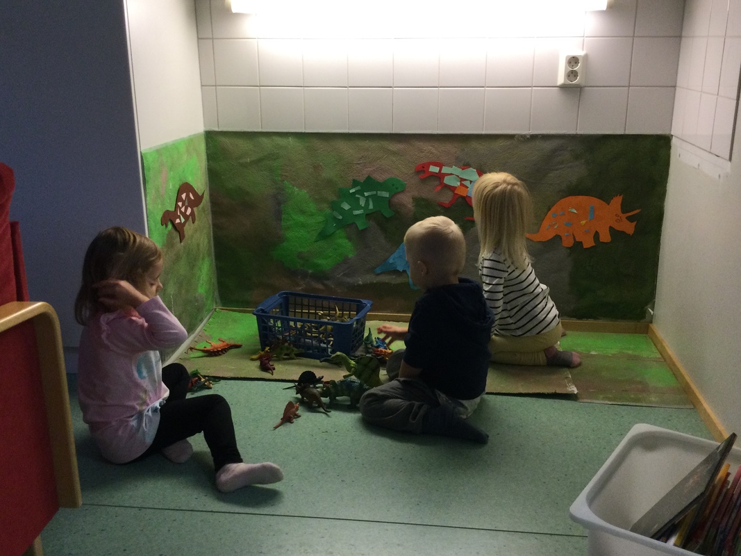 Kuvassa kolme lasta istuvat lattialla ja leikkivät dinosauruksilla