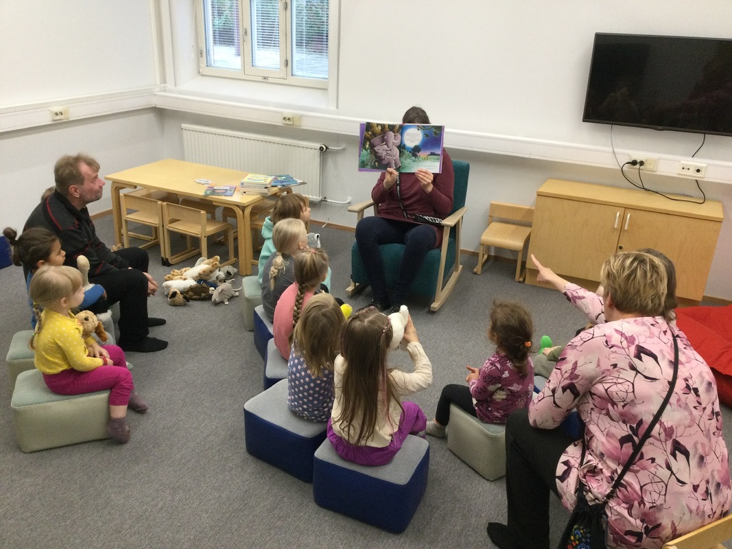 Lapset ja kaksi aikuista istuvat pehmojakkaroilla ja kuuntelevat kun yksi aikuinen lukee kirjaa