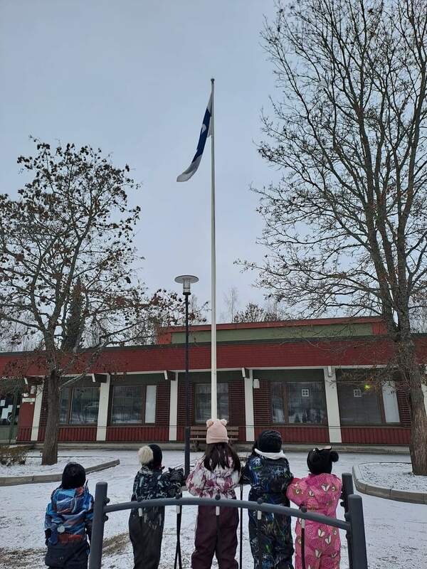 Lapset katsovat päiväkodin pihalla, kun Suomen lippu liehuu lipputangossa.