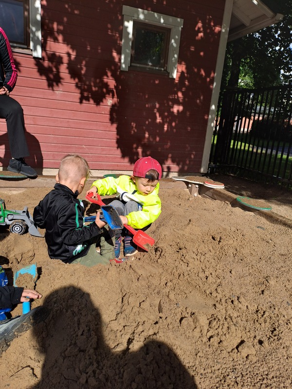 Kaksi lasta leikkii hiekkalaatikolla päiväkodin pihalla. Aurinko paistaa.