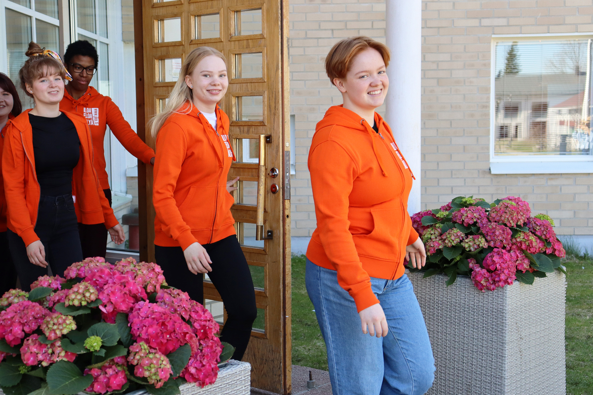 Iloiset opiskelijat kävelevät oppilaitoksen ovesta. Opiskelijoilla on päällä oransit hupparit. Oven vieressä kukkaistutukset.
