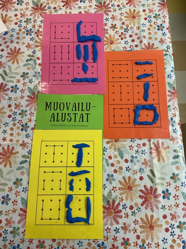 IMG_4175.jpeg Pöydällä pinkki, oranssi ja keltainen muovailualusta, joiden päälle on tehty sinisestä muovailuvahasta erilaisia kuvioita ja muotoja.