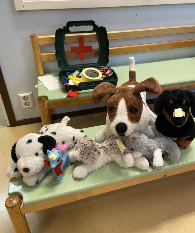 IMG_5092.jpeg Penkillä rivissä erilaisia pehmoleluja ja poneja, penkin takana lääkärinlaukku jossa lääkärileluja.