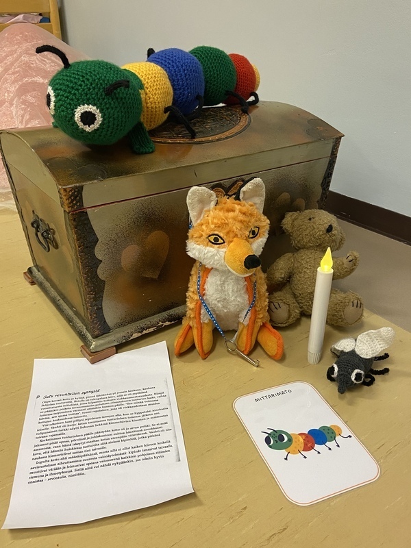 Laulutuokio ketun kanssa Pehmolelukettu ja -nalle istuvat aarrearkun edessä. Arkun päällä on virkattu mittarimatolelu. Lattialla on kuvakortti mittarimadosta ja ledkynttilä.