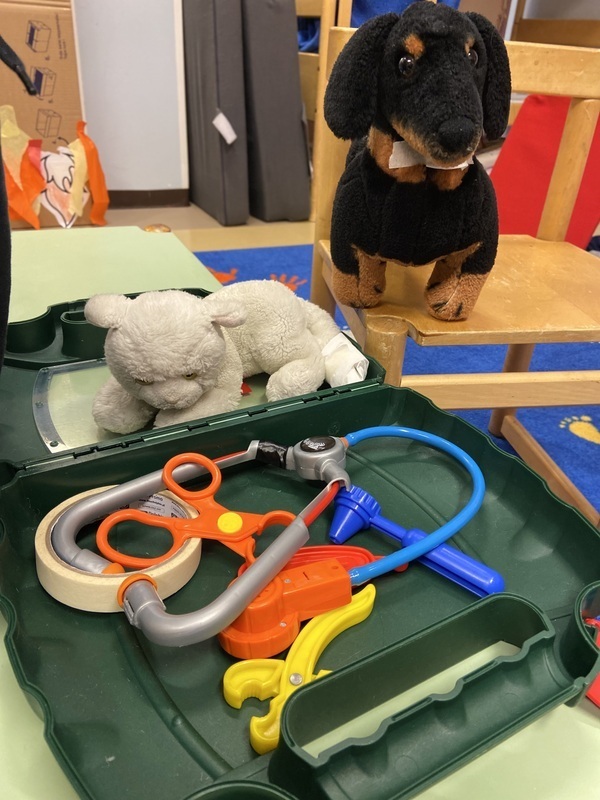 Kuva Lääkärilaukussa erilaisia lääkärin tarvikkeita mm. stetoskooppi, pehmolelukissalla paperinen side jalassa ja pehmokoira odottaa vuoroaan.