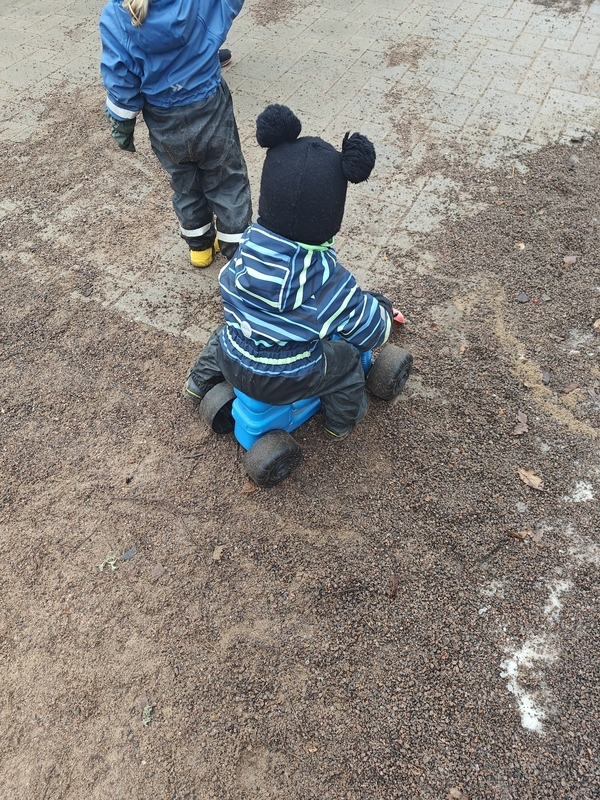 Kuvassa toinen lapsi istuu leikkimopon kyydissä, ja toinen seisoo vieressä.