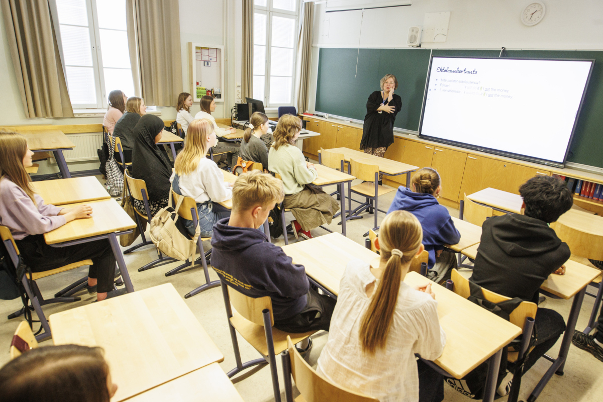 Koululaisia istumassa pulpeteissa luokkahuoneessa, taka-alalla seisoo opettaja.