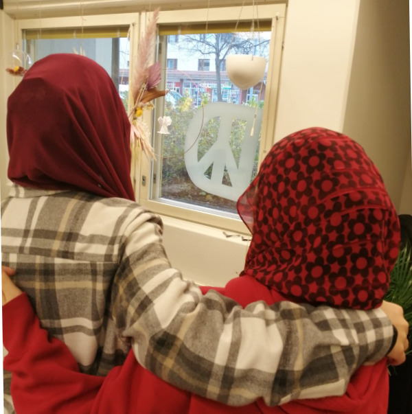 Kaksi huivipäistä tyttöä katselee ulos ikkunasta kädet toistensa olkapäillä. Ikkunassa näkyy askarreltu peace-merkki.
