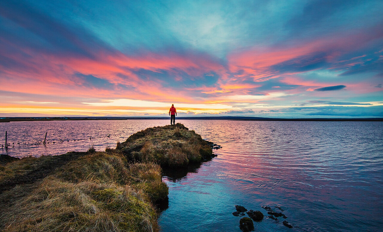 Ihminen seisoo rannalla ja katsoo kohti värikästä auringonlaskua.
