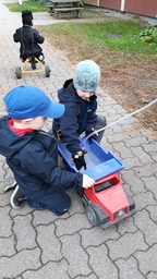 Kaksi lasta korjaavat yhdessä pihalla leikkikuorma-autoa. Yksi lapsi pyöräilee.