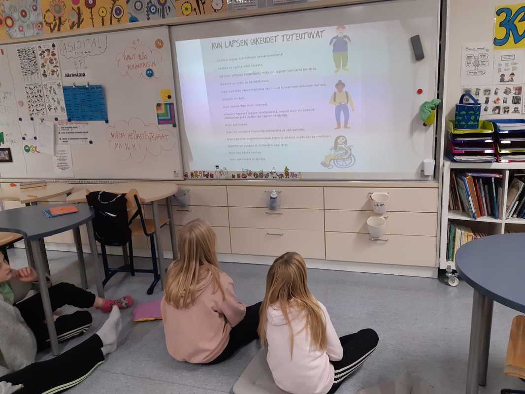 Oppilaat istuvat maassa ja seuraavat taululta lasten oikeuksien tietoiskua.