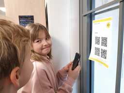 Tyttö katsoo toiseen oppilaaseen ja hymyilee. Tytöllä on kädessä iPad ja hän on ottamassa seinällä olevaa qr-koodia tähtäimeen.
