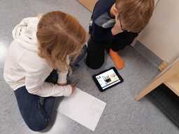 Oppilaspari istuu lattialla ja tekee tehtäviä iPadia hyödyntäen. Kuva otettu ylhäältä, kasvot eivät näy.