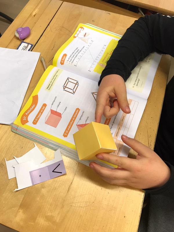 Pulpetti, jolla on matematiikan oppikirja auki kuution opetussivulta, oppilaan kädet näkyvät: oppilas rakentaa kuutiota pahvista ohjeiden mukaan.
