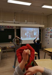 Kuva otettu luokan takaa, oppilaat ovat selin ja tanssivat taululle heijastetun videon mukaan.