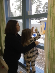 Lapsi ja aikuinen seisovat ikkunan edessä ja kiinnittävät lumihiutaleaskartelua ikkunaan.