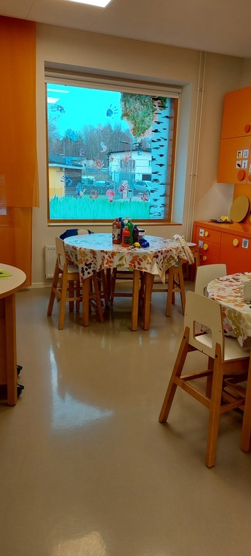 Pöydät joissa liinat, oranssit kalusteet, ikkunaan on tehty lasten kädenjälkiä maalaamalla.