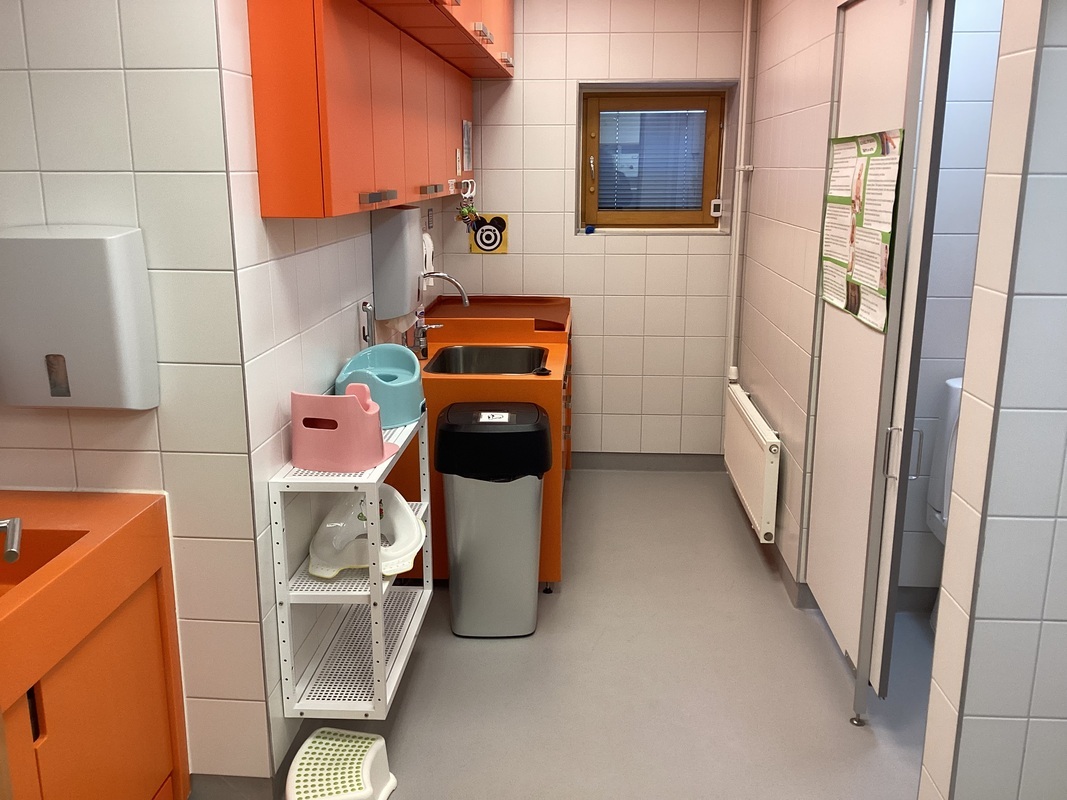 Vasemmalla oven takana käsienpesupaikka. WC:n takaosassa vaipanvaihtopaikka. Oikealla kaksi wc pönttöä ja pottia.
