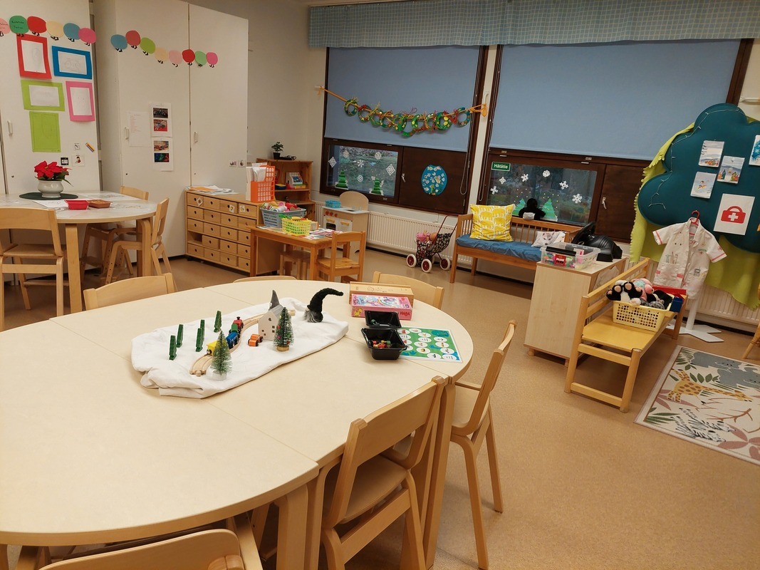 Iso pöytä keskellä. Ympärillä erilaisia leikkipaikkoja. Takana ikkunat, joista voi katsoa ulos.