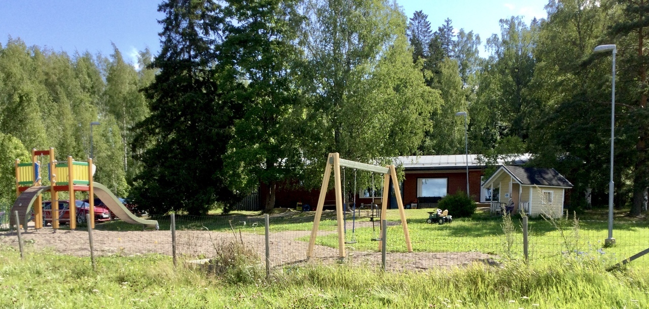 Koisjärven päiväkodin piha-aluetta ja taka-alalla päiväkodin rakennus. Rakennuksen edessä näkyy keinut, karuselli, leikkimökki ja kiipeilyteline, jossa liukumäki. Piha-alueella puita, nurmikkoa ja hiekka-alue.