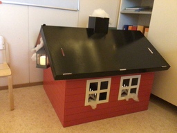 Punainen pahvilaatikko mökki, jossa koristeena jouluvaloja.