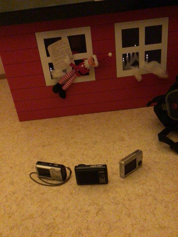 Minni-tonttu roikkuu pahvimökin ikkunasta kädessään lappu ja edessä maassa on kolme kameraa.