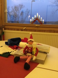 Minni-tonttu istuu pöydällä. Sylissään sakset sekä liimapuikko ja ympärillään muita askartelutarvikkeita.