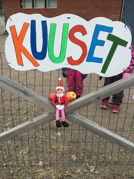 Minni-tonttu istuu päiväkodin aidan päällä, vieressään kaksi omenaa. Yläpuolella kyltti, jossa lukee kuuset.