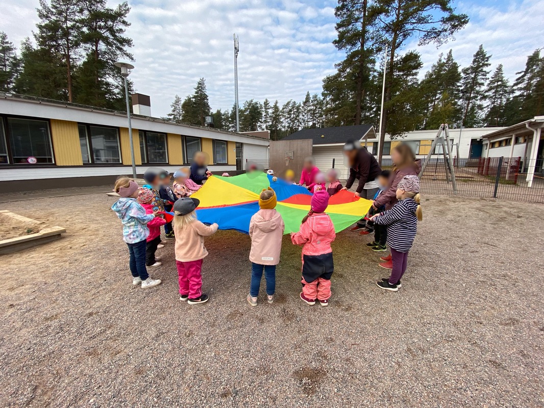 Leikkivarjolla jumppaamista ja hauskanpitoa pihalla. Useammasta ryhmästä tuli lapsia ja aikuisia leikkimään leikkivarjolla. Pidimme kaikki varjon reunoista kiinni ja heiluttelimme varjoa.