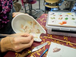 Kuvan vasemmassa reunassa oleva henkilö pitää kädessään valmisteilla olevaa ruskeansävyistä posliininmaalaustyötä ja maalaa pensselillä kuviota näkyväksi. Pöydällä on vahakangasliina ja maalaustarvikkeita.
