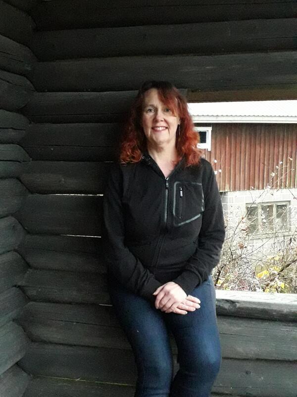 Kuvassa nainen istuu pihakatoksessa. Taustalla vanha navetta.